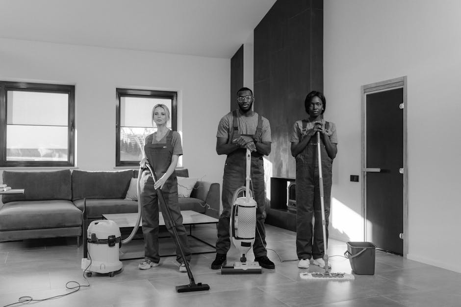Three professional cleaners from Cleaner Ilford are performing surface cleaning in a modern living room. The team includes a woman on the left using a vacuum cleaner on the tiled floor, a man in the center holding a spray bottle and standing behind an upright vacuum with a corded power supply, and a woman on the right using a mop and bucket for floor mopping. The room features a light-colored tiled floor, a large dark wooden accent wall, and two windows allowing natural light to brighten the space. A sofa with cushions is visible against the left wall, and a small cabinet is positioned beneath the windows. The cleaning tools and equipment suggest thorough domestic cleaning, emphasizing hygiene and maintenance as part of deep cleaning services by Cleaner Ilford, highlighting their commitment to avoiding hidden charges in Ilford cleaning quotes.