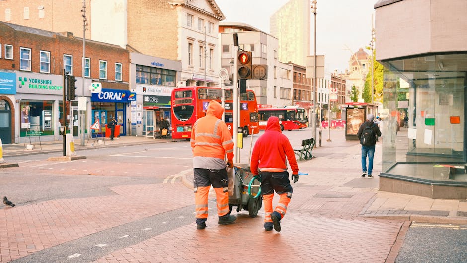 Two street cleaners dressed in orange high-visibility uniforms are performing surface cleaning tasks on a sidewalk in Ilford, near some shop fronts and a bus stop. They are pushing a small cleaning trolley with cleaning tools, maintaining the cleanliness of the paved pathway. The background features a red double-decker bus, various retail stores, and pedestrians walking along the street. The area is well-lit with natural daylight, and the storefronts, including a glass-fronted shop, are clean and orderly. The scene emphasizes routine urban maintenance carried out by professional cleaning services to ensure hygiene and a tidy environment, exemplifying the type of service provided by Cleaner Ilford linked to local commercial cleaning and maintenance.