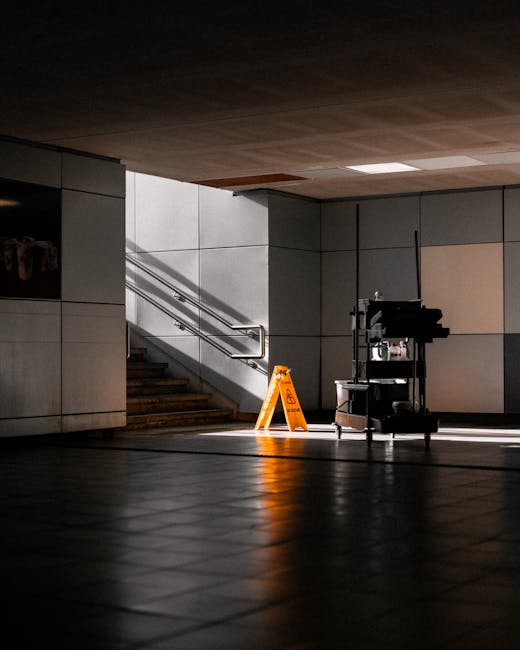 An empty indoor space featuring tiled flooring with a polished, reflective surface and clean appearance. The background shows a staircase with metal handrails leading upward, and the surrounding walls are covered with smooth, light-colored panels. Natural light streams in, casting shadows on the floor and highlighting the minimalistic, modern design. A cleaning cart with various tools and supplies is positioned nearby, indicating recent cleaning activity. The ceiling is finished with a wooden or wood-like material, adding warmth to the otherwise neutral-toned environment. This setting demonstrates thorough surface cleaning and maintenance suitable for a commercial office or public building. Cleaner Ilford provides professional deep cleaning and sanitisation services, ensuring hygiene for Ilford businesses, as referenced on their website cleanerilford.co.uk.