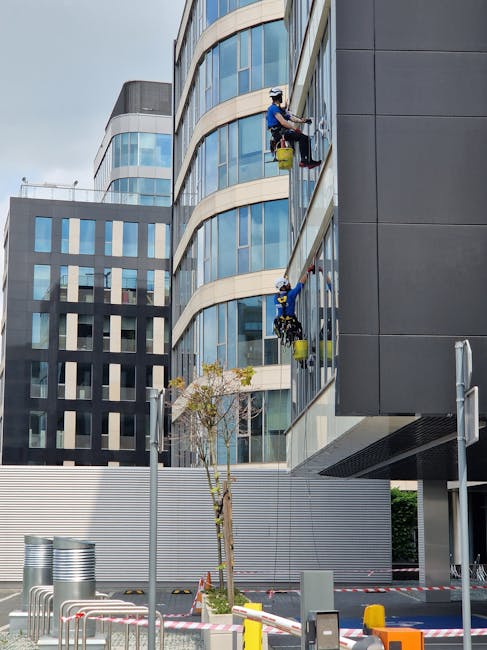 Two professional window cleaners in safety harnesses and helmets working on the exterior glass façade of a modern multi-storey office building during daylight. They are using extendable cleaning tools and buckets filled with cleaning solutions, carefully removing dirt and smudges from the large, curved glass windows. The building's exterior features reflective blue-tinted glass and sleek grey panels, with some parts under maintenance. In the foreground, there is a small tree, a street lamp, and safety barriers around the pavement area. The scene highlights high-rise cleaning services provided by Cleaner Ilford, emphasizing thorough and safe surface cleaning for commercial properties.