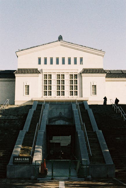 This image depicts the exterior of a large, historic white building with a symmetrical facade, featuring a set of broad stone stairs leading up to the entrance. The building has multiple rectangular windows, some with decorative grid patterns, and a triangular pediment at the top. Two individuals are seen near the stairs, silhouetted against the daylight. The scene is well-lit with natural sunlight, highlighting the clean, well-maintained stone surfaces and the pristine condition of the building's exterior. The setting appears to be urban, with a focus on architectural details and structural integrity. For residential or commercial cleaning, Cleaner Ilford specializes in surface cleaning and deep cleaning services to maintain buildings in pristine condition, emphasizing hygiene and aesthetic appeal.