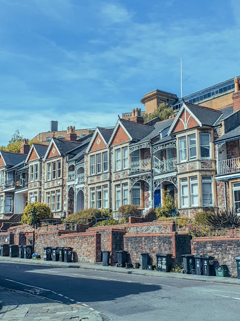 A row of Victorian-style terraced houses with decorative white trim and bay windows, situated along a residential street under a bright blue sky. The houses are built with red brick and feature small front gardens with steps leading up to the entrances. The street is lined with black wheelie bins for waste collection, and the area appears well-maintained and clean, reflecting a typical suburban neighbourhood. This image showcases the exterior of residential buildings that could benefit from professional cleaning services provided by Cleaner Ilford, emphasizing the importance of surface cleaning and property maintenance in domestic settings.