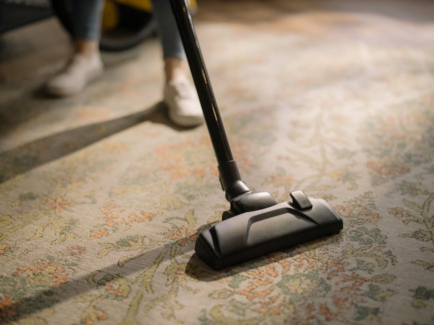Close-up view of a vacuum cleaner with a transparent dust container filled with debris, connected to a flexible, striped hose attached to a floor brush. The vacuum is placed on a pinkish-beige carpet