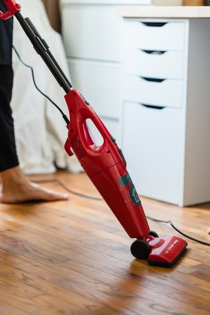 Close-up of a red cordless vacuum cleaner being used on a wooden floor in a domestic setting, with a person barefoot nearby. The vacuum is positioned upright, actively cleaning the polished hardwood surface, which appears dust-free and shiny. To the right, a white storage unit with multiple drawers is visible, indicating a tidy and well-maintained room. The lighting highlights the clean, smooth texture of the wooden flooring, emphasizing surface cleaning and hygiene. This scene reflects professional domestic cleaning performed by Cleaner Ilford, showcasing tools and surfaces typical of a home environment associated with routine deep cleaning and surface sanitisation.