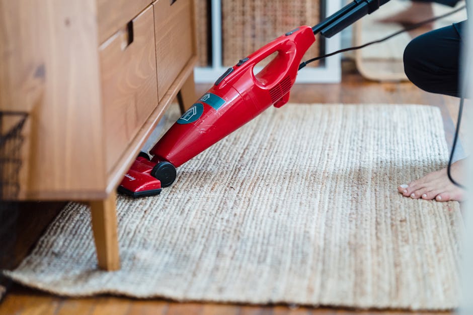 Close-up of a red cordless vacuum cleaner being used on a wooden floor in a domestic setting, with a person barefoot nearby. The vacuum is positioned upright, actively cleaning the polished hardwood surface, which appears dust-free and shiny. To the right, a white storage unit with multiple drawers is visible, indicating a tidy and well-maintained room. The lighting highlights the clean, smooth texture of the wooden flooring, emphasizing surface cleaning and hygiene. This scene reflects professional domestic cleaning performed by Cleaner Ilford, showcasing tools and surfaces typical of a home environment associated with routine deep cleaning and surface sanitisation.