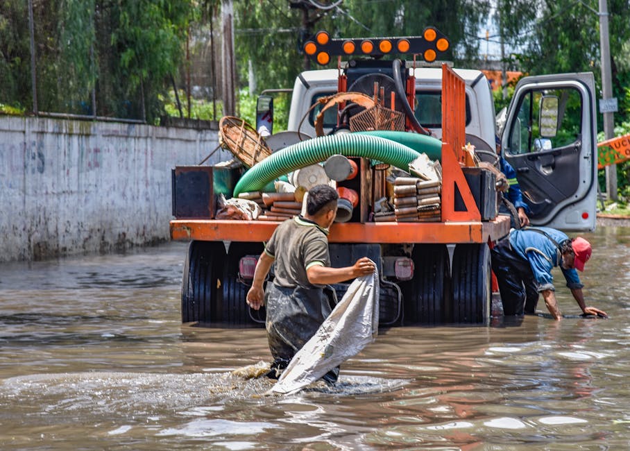 Two workers in waterproof clothing are engaged in emergency flood cleaning near a large orange truck filled with equipment and supplies, which is parked in a flooded outdoor area with water reaching u